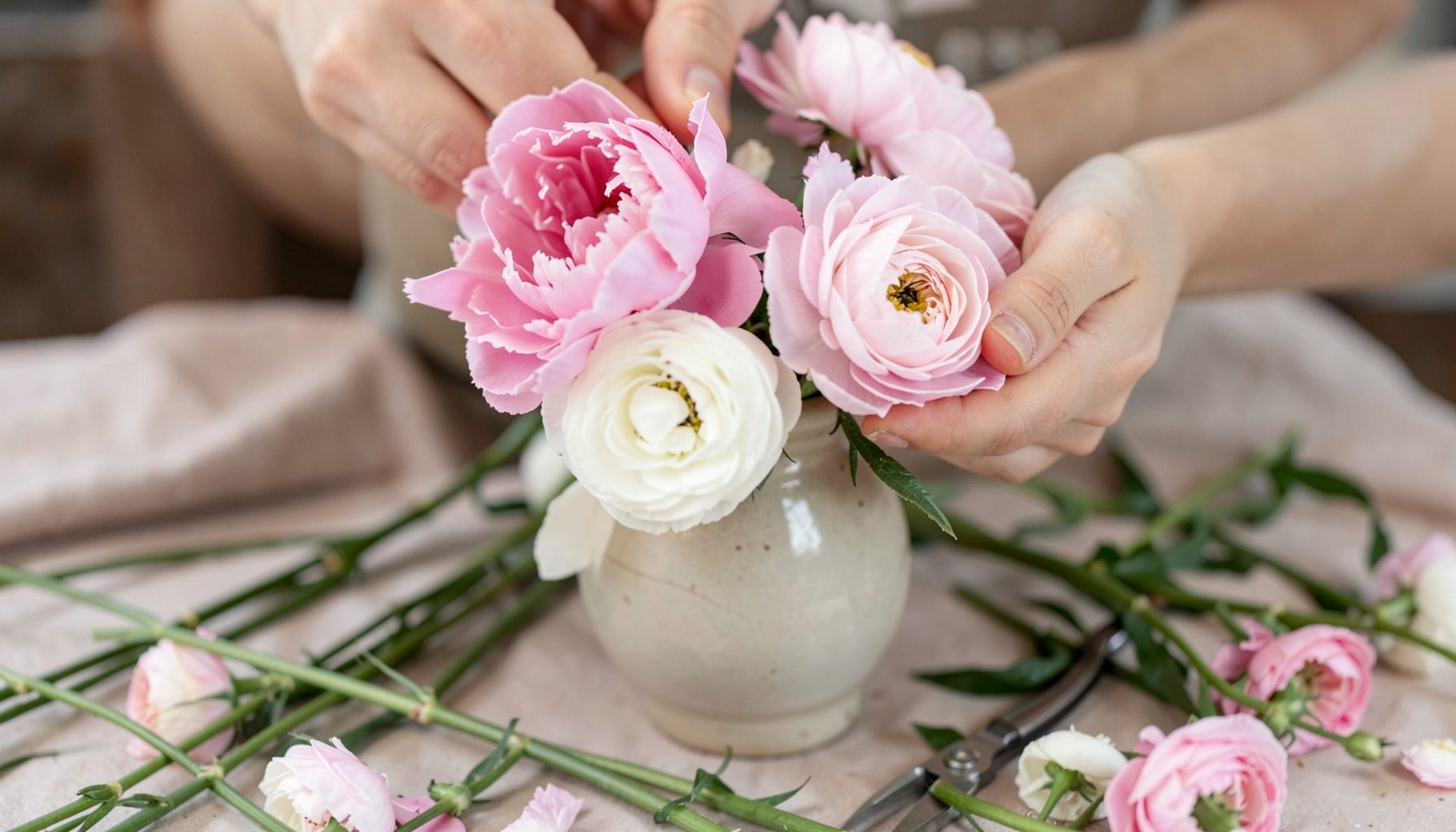 A close-up of hands arranging pink peonies and white ranunculus in a ceramic vase, surrounded by loose stems and floral shears