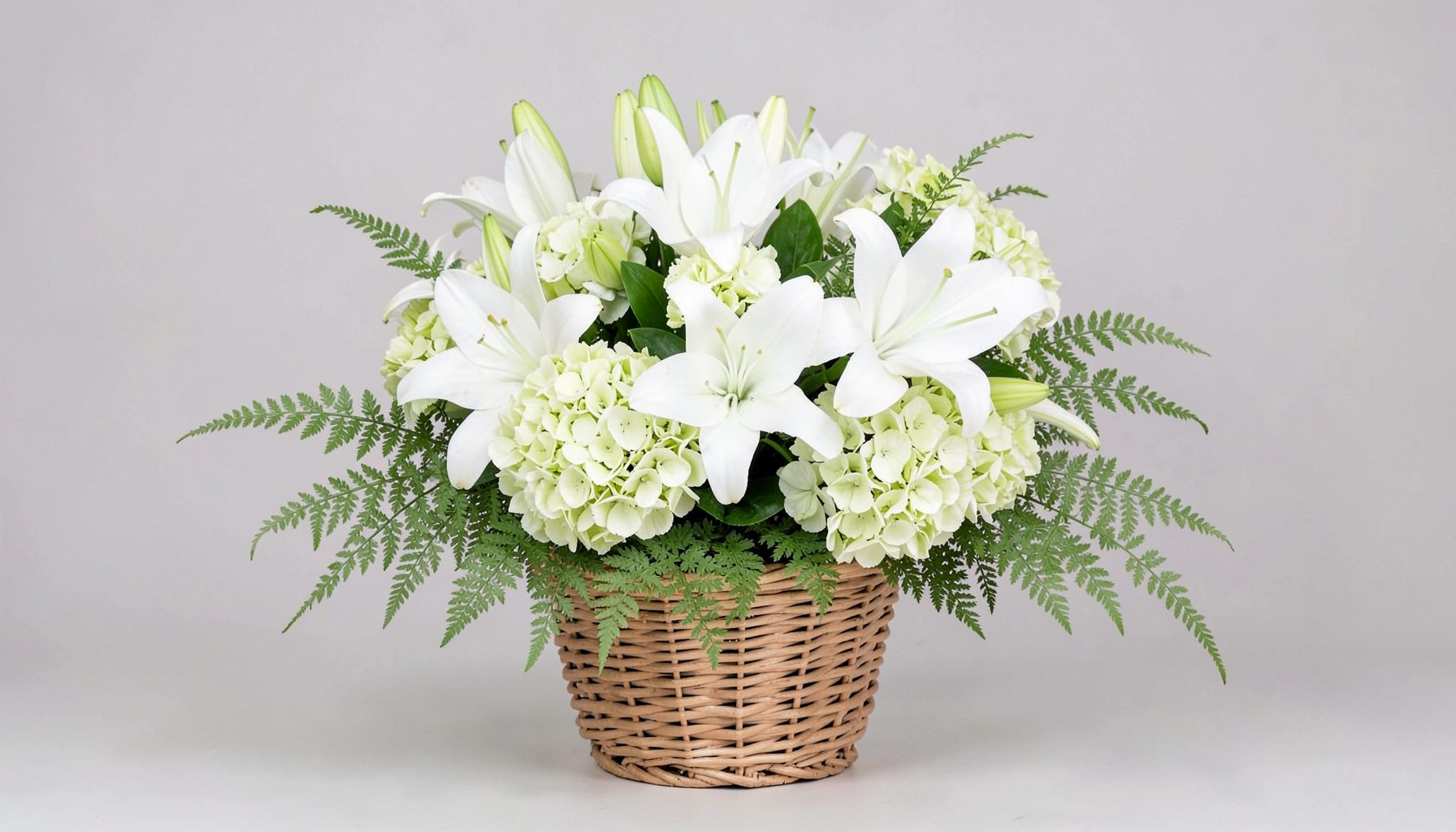 A sympathy arrangement of white lilies, pale green hydrangeas, and soft ferns in a woven basket against a neutral background