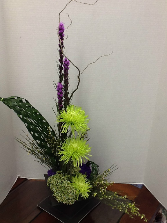 Modern arrangement with green spider mums, purple flowers, curly willow branches, and a woven leaf accent on a dark stand