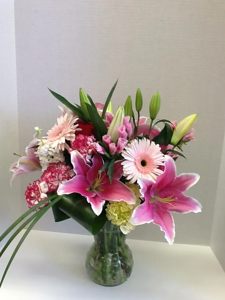 A glass vase holding a pink and white bouquet with stargazer lilies, gerbera daisies, carnations, and greenery against a light background