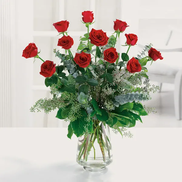 A glass vase filled with a dozen red roses and green eucalyptus foliage, set on a white surface against a light, airy interior