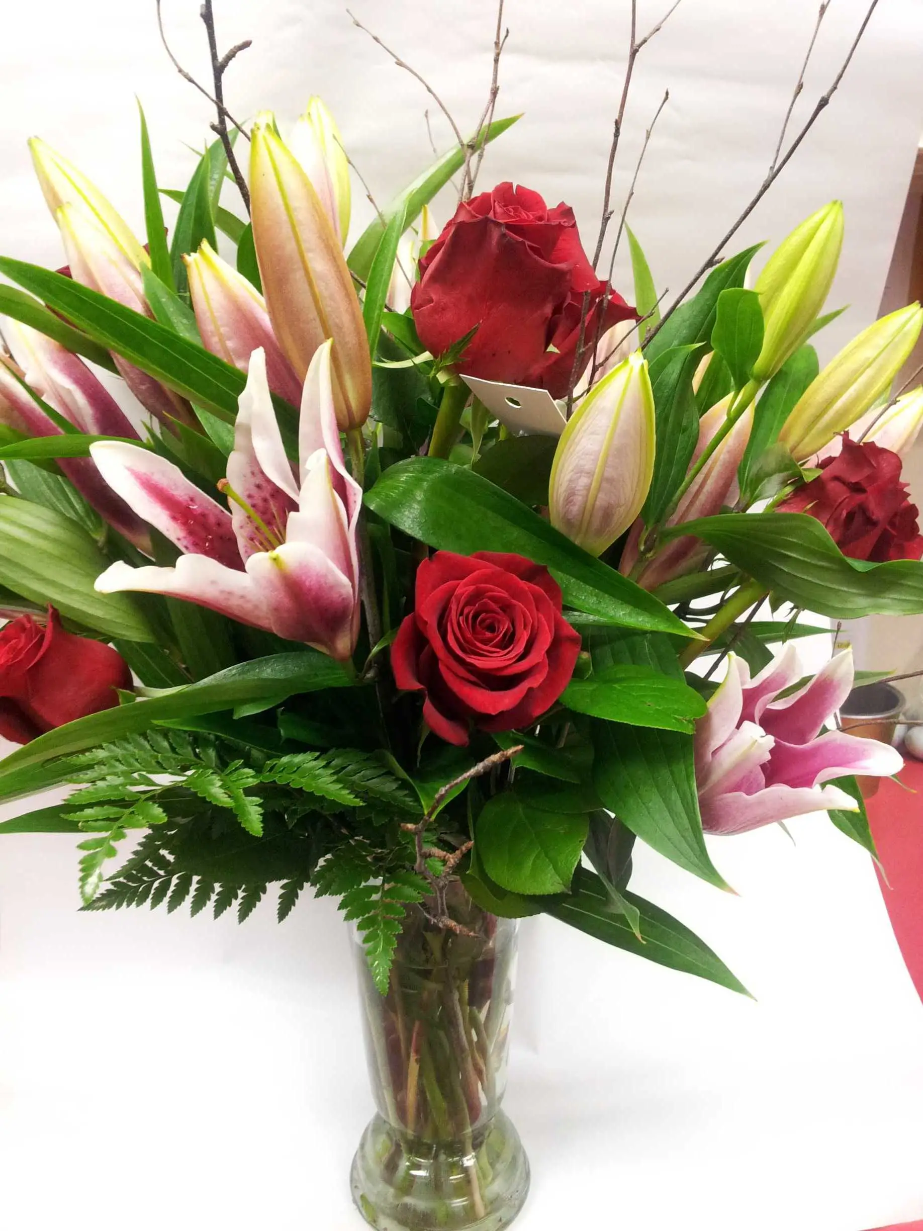 A close-up bouquet of red roses and pink stargazer lilies with green foliage and fern in a clear glass vase against a pale background