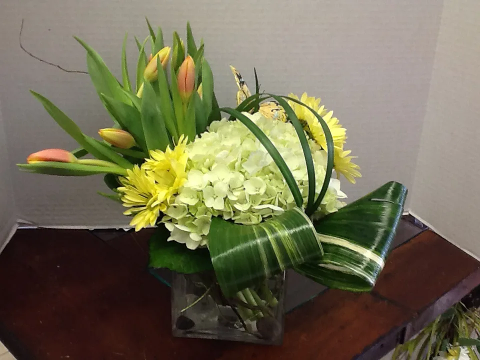 Floral arrangement with yellow daisies, green hydrangea, orange tulips, and decorative leaf swirl in a glass cube vase on a white surface